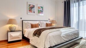Un dormitorio moderno con una cama grande, dos mesitas de noche, una mesilla auxiliar, una ventana con cortinas translúcidas y dos cuadros con motivos florales en la pared.