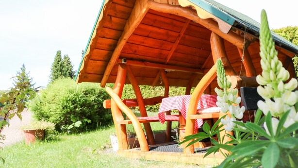 Eine Holzhütte mit einem geneigten Dach, einer Bank und einem Tisch mit karierter Tischdecke.
