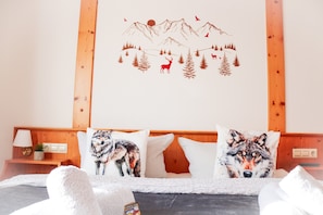 A bedroom with a mountain-themed wall decal, two wolf pillows, a bed with white bedding, a nightstand with a lamp and a plant, and wooden paneling. - Wise Wolf – Standard Double Room at House of Happiness with Sunny Balcony (Schluchsee)