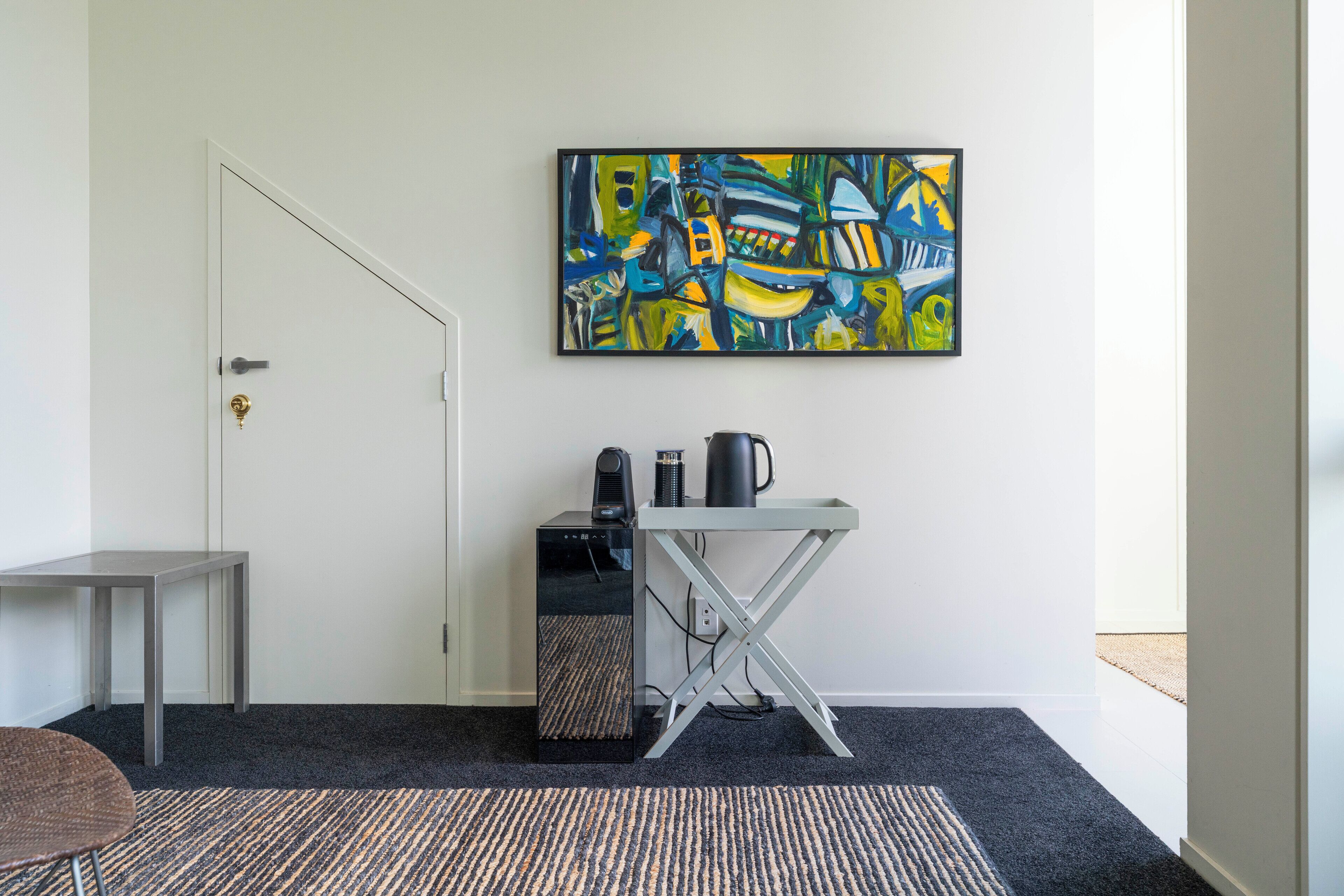 A room with a white door, a small table, a desk with a computer and a coffee maker, and a colorful abstract painting on the wall.
