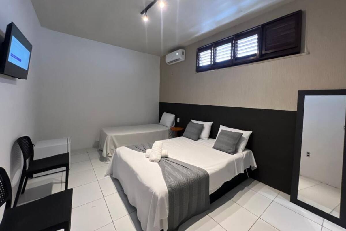 A hotel room with a bed, a desk, a chair, a TV, and an air conditioning unit.