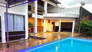A swimming pool with a tiled deck, a covered seating area, and a stone wall in the background. - Pousada Jardins (São Luís)