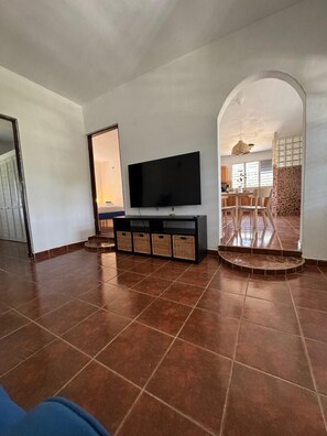 A living room with a flat-screen TV on a black entertainment unit, a dining area with a table and chairs, and a kitchen visible through an archway. - Casa Aurora - Family 3BR Near Ferry / AC, WiFi & Laundry (Vieques)