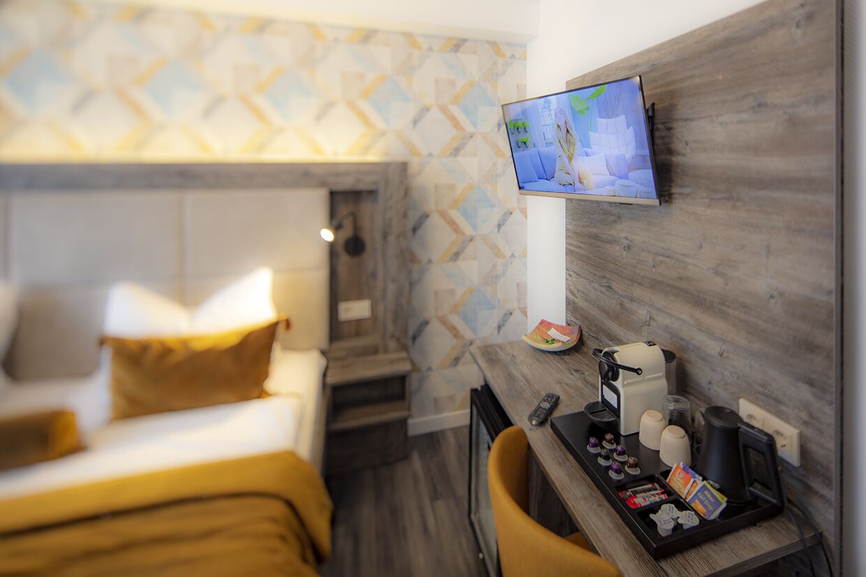 Hotelkamer met een bed, een bureau met koffiezetapparaat, een aan de muur bevestigde televisie en een behang met patroon.