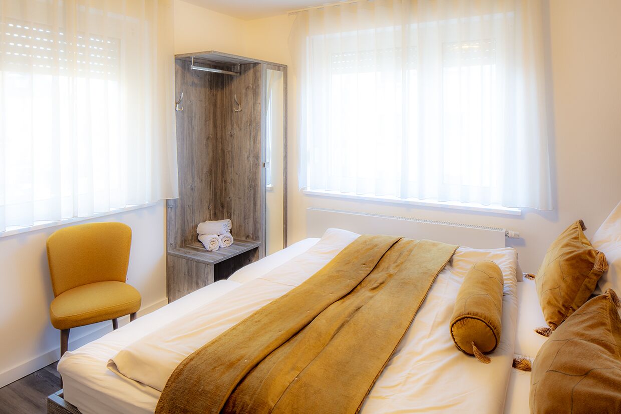 A hotel room with a bed, a wardrobe, a chair, and a window with sheer curtains.
