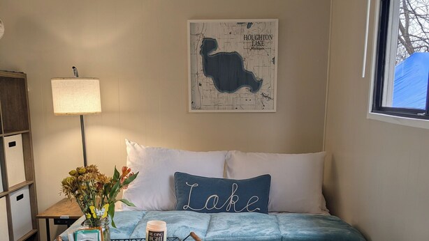 Ein Schlafzimmer mit einem Bett, Kissen, einer Karte an der Wand, einer Lampe, einer Vase mit Blumen und einem Fenster.