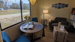 Ein gemütliches Zimmer mit einem großen Fenster, einem Tisch mit Buch und Blumenvase, einer Couch, einer Lampe und einem Koffer.