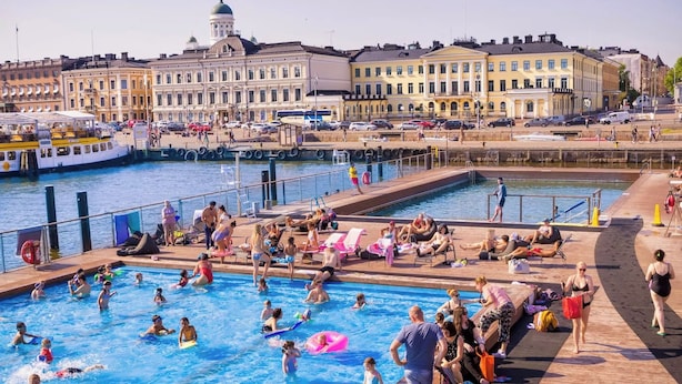 A public swimming pool with people swimming and sunbathing, surrounded by a waterfront promenade with buildings and a boat docked nearby. - Superior Suite by the Sea and Market Square (Helsinki)