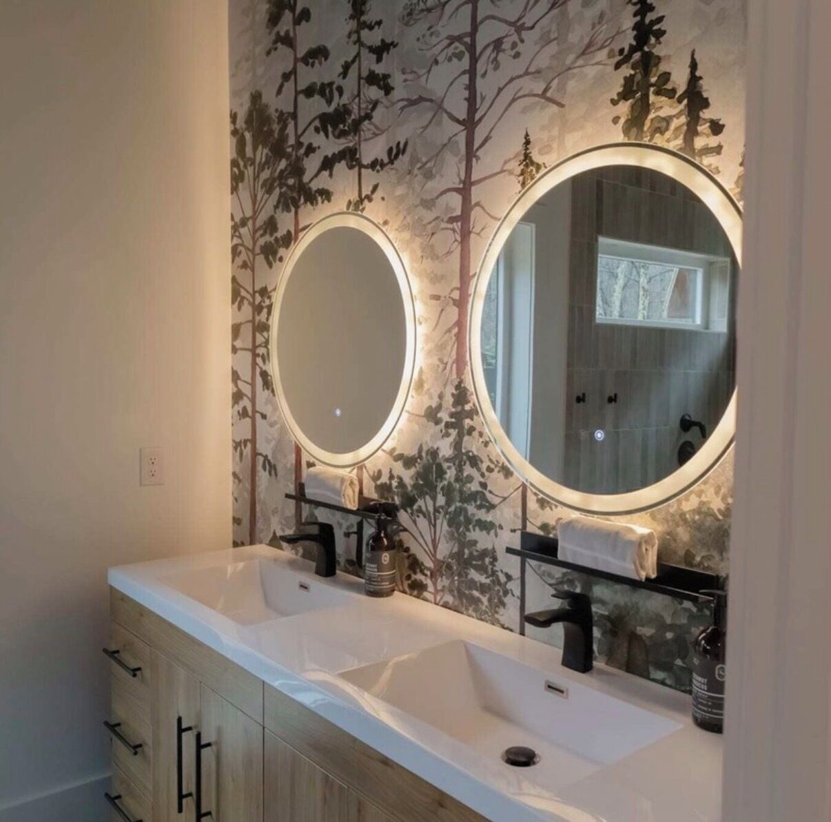 Une salle de bain avec deux lavabos, un grand miroir et un mur décoré d’un motif représentant un arbre et une forêt.