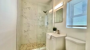 Ein Badezimmer mit einer gläsernen Duschkabine, weißen Marmorfliesen, einer weißen Toilette und einem Fenster mit Jalousien.