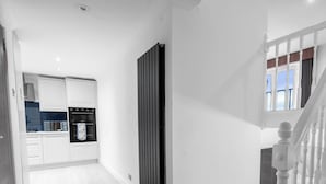 Una cocina moderna con armarios blancos y electrodomésticos integrados, un radiador negro vertical y una escalera con barandilla blanca y escalones alfombrados en marrón.