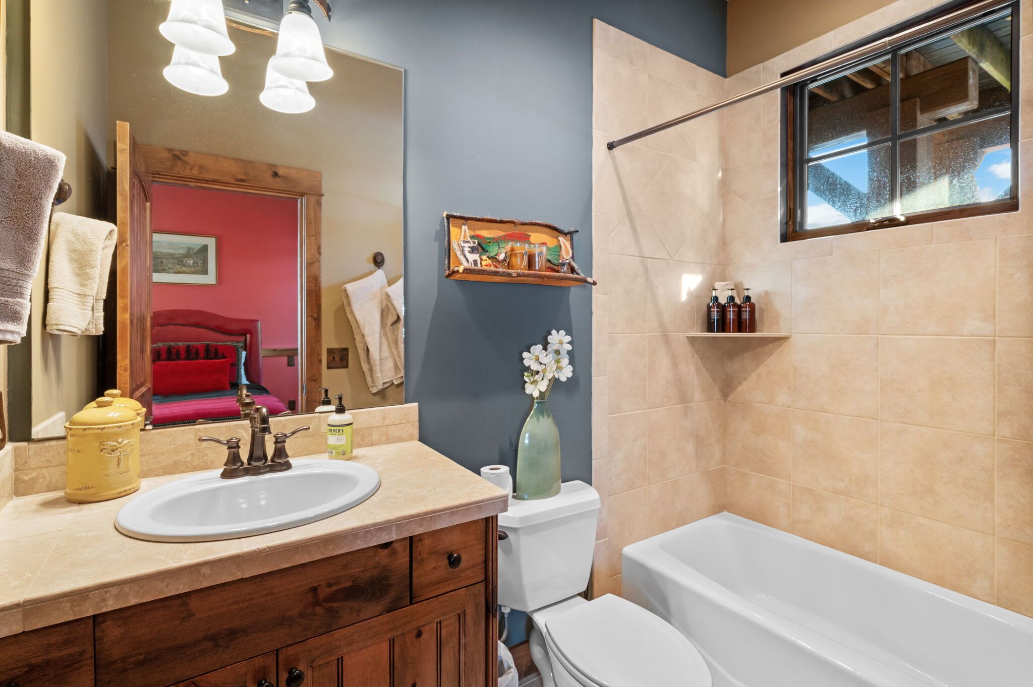 Un baño con lavamanos, inodoro y bañera. Hay un espejo, un perchero para toallas y un estante con artículos de aseo.