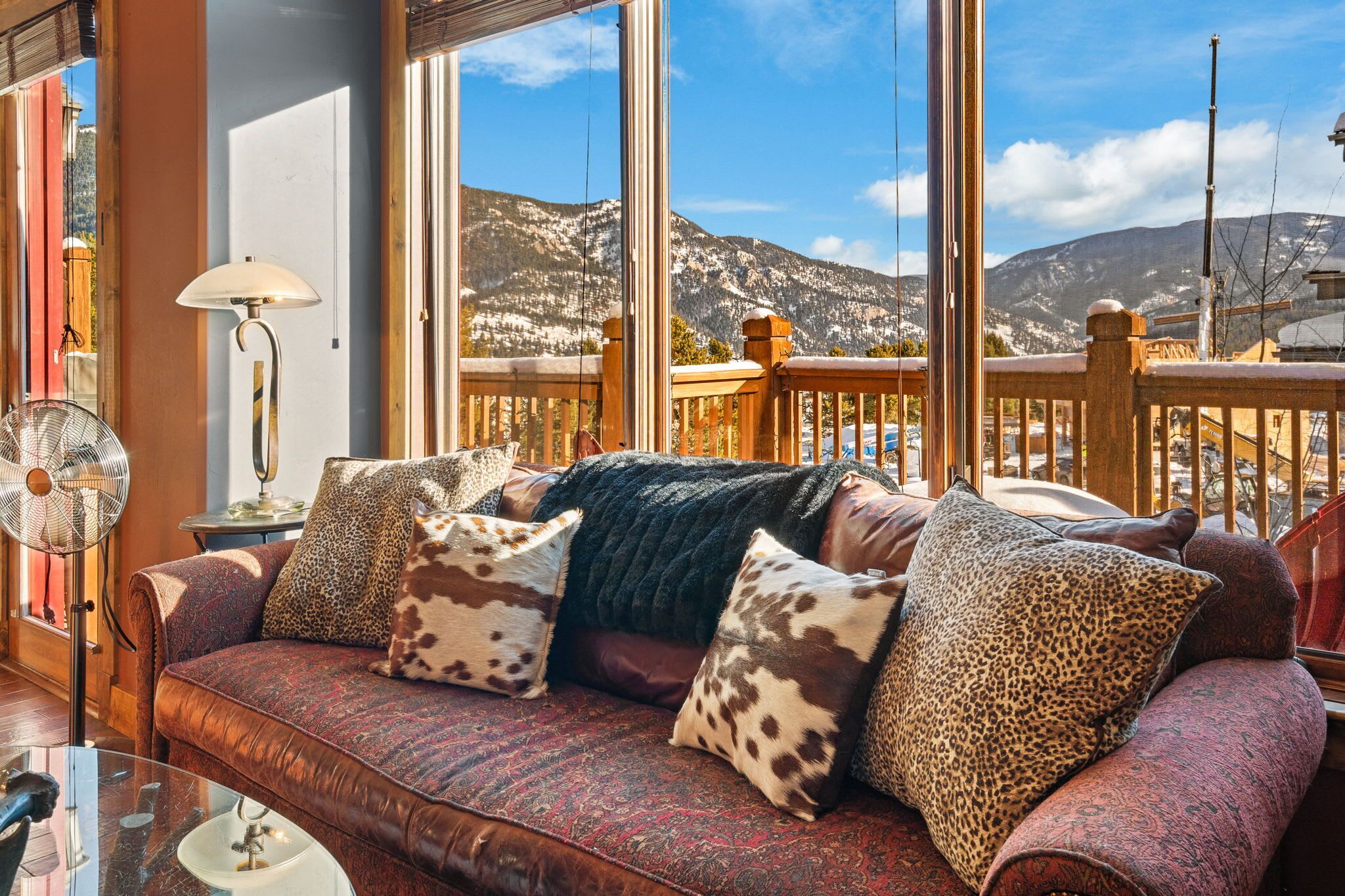 A cozy living room with a large window offering a mountain view, a wooden deck, and a leather sofa with patterned cushions.