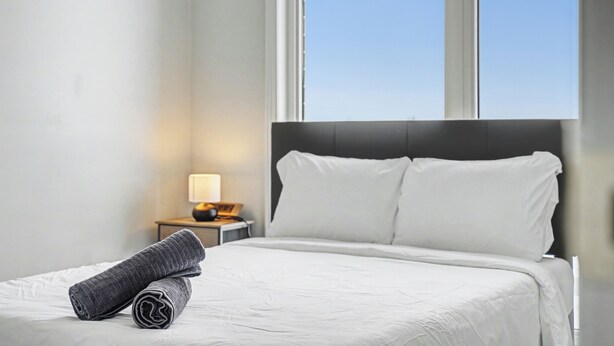 A neatly made bed with white linens, a grey towel, and a nightstand with a lamp. - Cedar Nest - 5 Beds, 3.5 Baths, 2 Park, Backyard (Pickering)