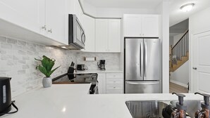 A modern kitchen with white cabinets, a stainless steel refrigerator, a coffee maker, and a potted plant. - Cedar Nest - 5 Beds, 3.5 Baths, 2 Park, Backyard (Pickering)