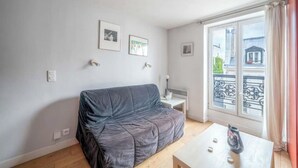 Una sala de estar con un sofá gris, una mesita blanca y una ventana con vistas a edificios.