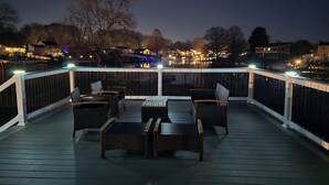 A wooden deck with wicker furniture and a railing, overlooking a body of water with buildings in the background. - Rumsey Island! (Joppatowne)