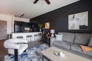 A modern living room with a grey sofa, a white armchair, a wooden coffee table, and a kitchen in the background.