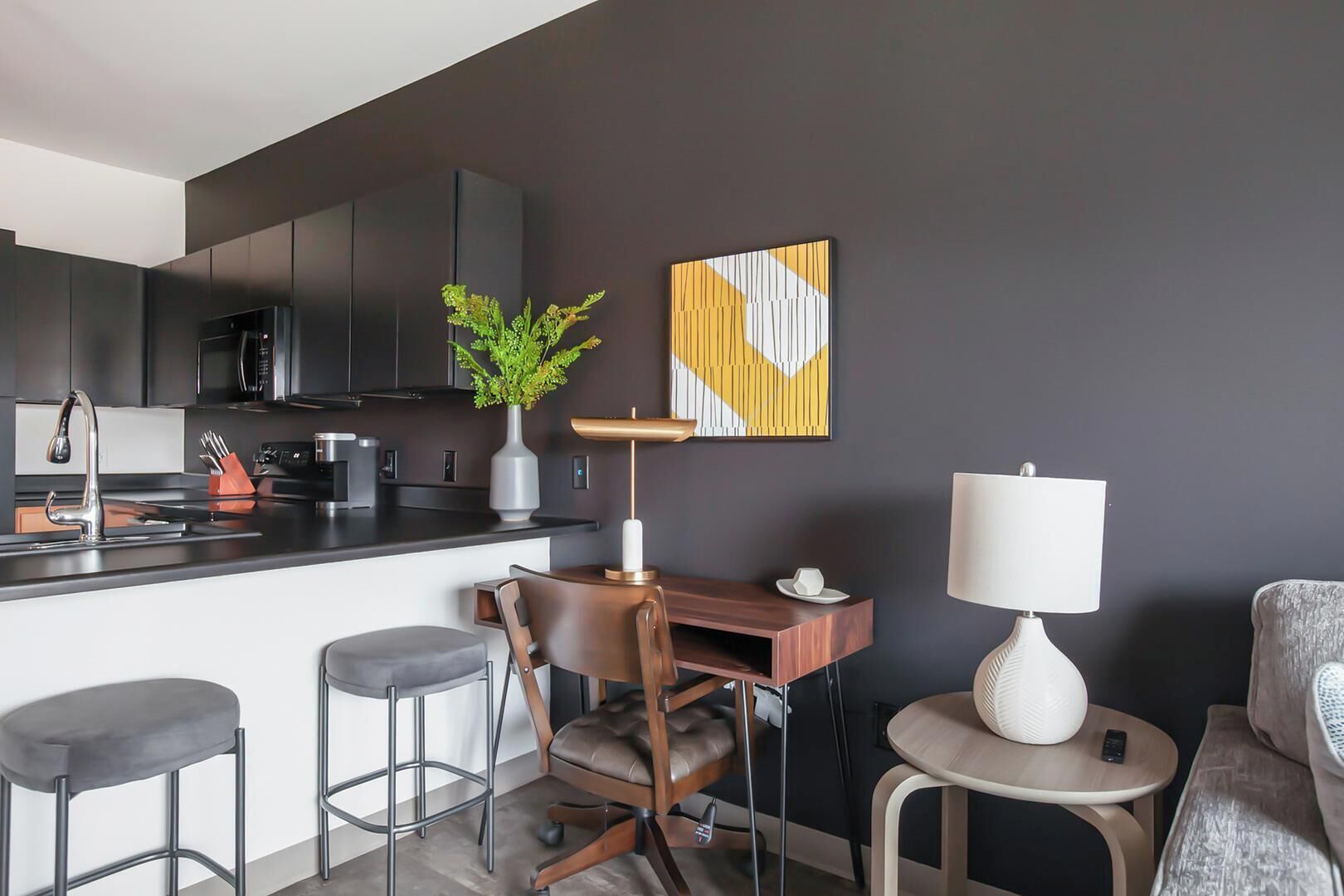 Une cuisine moderne avec des meubles de couleur sombre, un îlot central, des tabourets de bar, une chaise en bois, une petite table avec une lampe et une plante décorative.