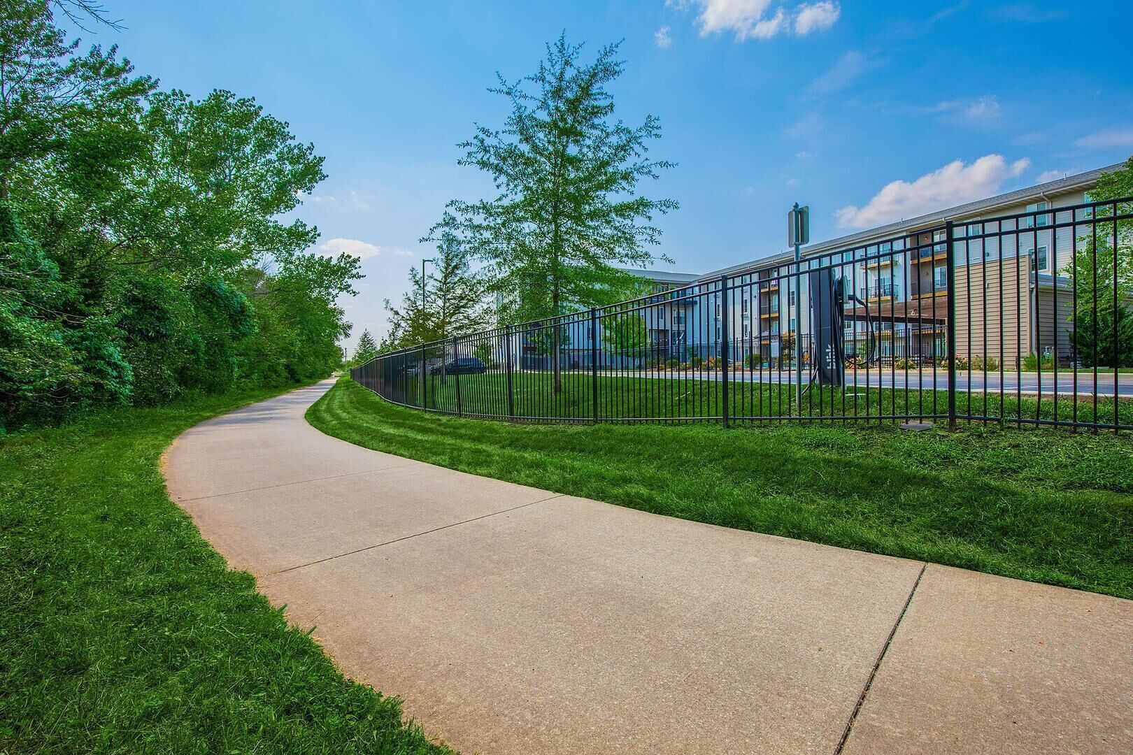 A curved pathway with a metal fence, surrounded by green grass and trees, leading to a building with a modern design.