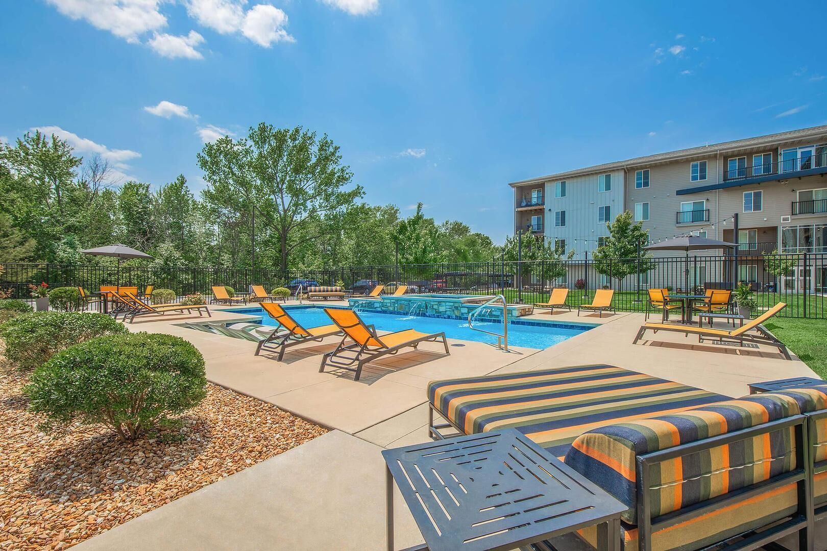 A pool area with lounge chairs and a small table, surrounded by a fence and trees.
