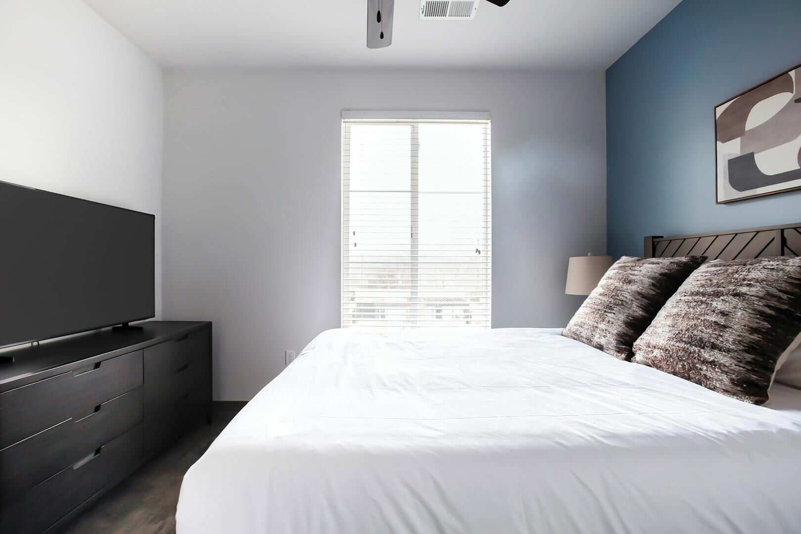 A modern bedroom with a large bed, a TV on a dresser, and a window with blinds.
