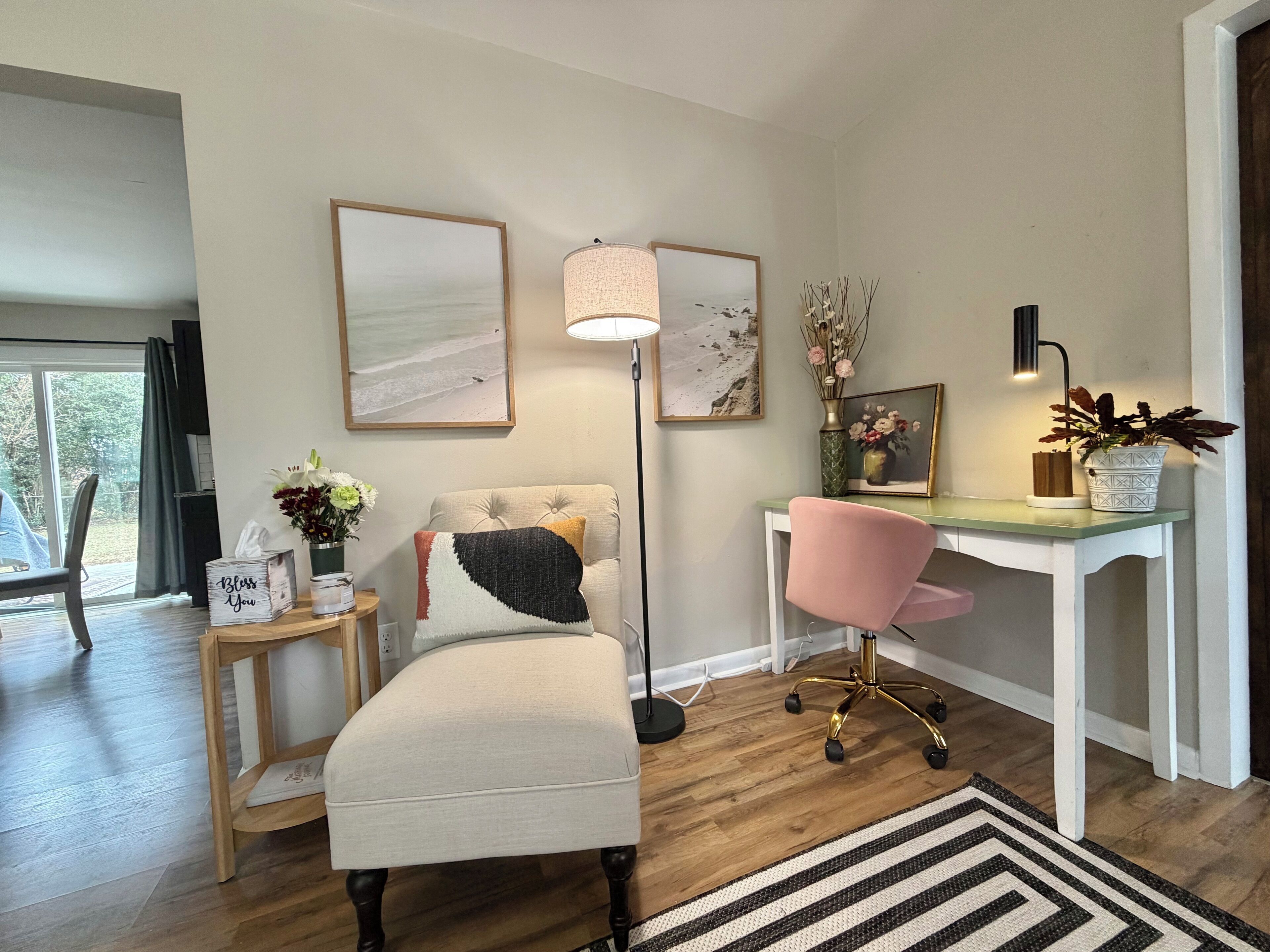 A modern living room with a sofa, a desk, and a chair. There are two framed pictures on the wall, a floor lamp, and a vase with flowers.