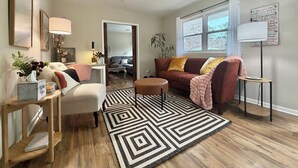 Una sala de estar con ventilador de techo, alfombra estampada, sofá, sillón, mesita y lámpara.