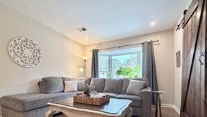 Un salón con un sofá seccional gris, una mesa de centro de madera, un adorno de pared, una lámpara de pie y una ventana con cortinas.