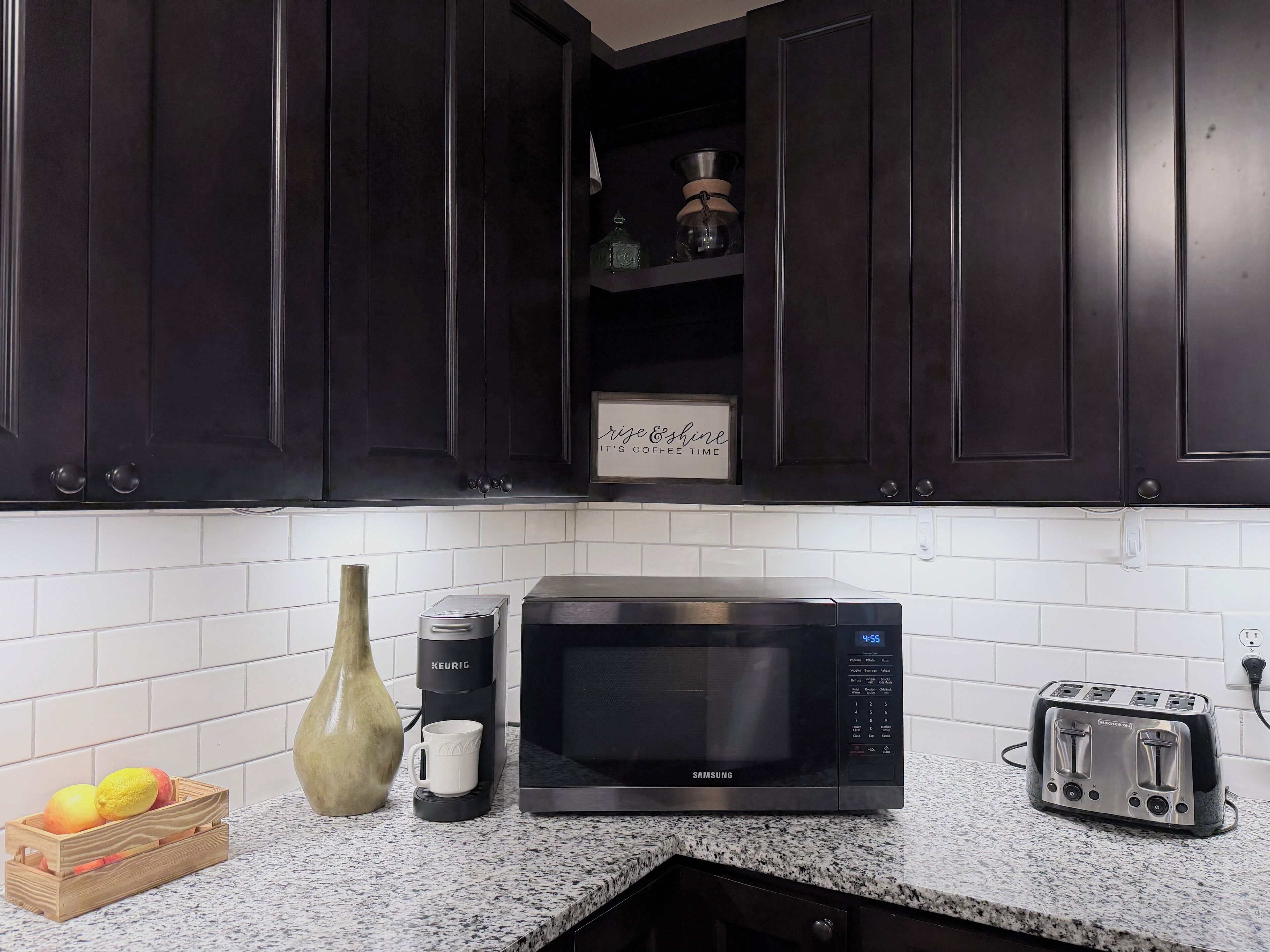 A kitchen with dark cabinets, a microwave, a toaster, a coffee maker, and a fruit basket.