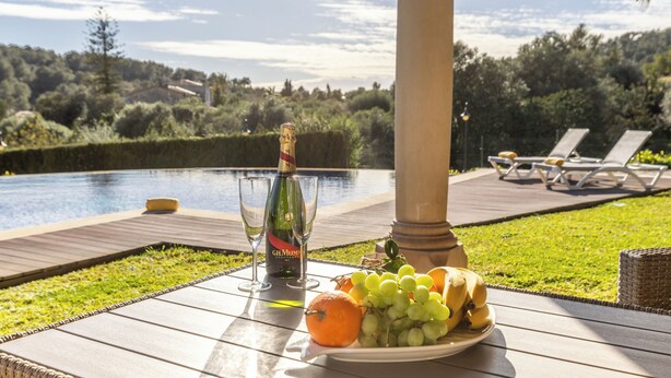 Una mesa con una botella de champán, copas y un plato de fruta junto a una piscina, con sillones de descanso al fondo.