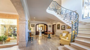 Un vestíbulo espacioso con piso de mármol, una imponente escalera con barandales de metal ornamentados y un sillón amarillo.