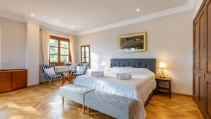 Un dormitorio con una cama grande, una zona de estar con sillas, suelo de madera y una ventana con cortinas.