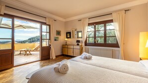 Habitación de hotel con dos camas, suelo de madera, balcón con mesa y sillas y vistas a las colinas.