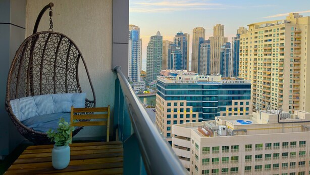 A balcony with a wicker swing chair, a small wooden table with a potted plant, and a view of a cityscape with high-rise buildings. - 35 OFF in Top View Studio in Marina (Dubai)