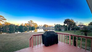 A wooden deck with a barbecue grill, overlooking a lake with trees and a distant building. - Grand Lagoon Retreat | Blue Angels & Boat Parking (Pensacola)