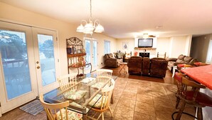 A spacious living area with a glass dining table, cushioned chairs, a fireplace, a television, and a large window. - Grand Lagoon Retreat | Blue Angels & Boat Parking (Pensacola)