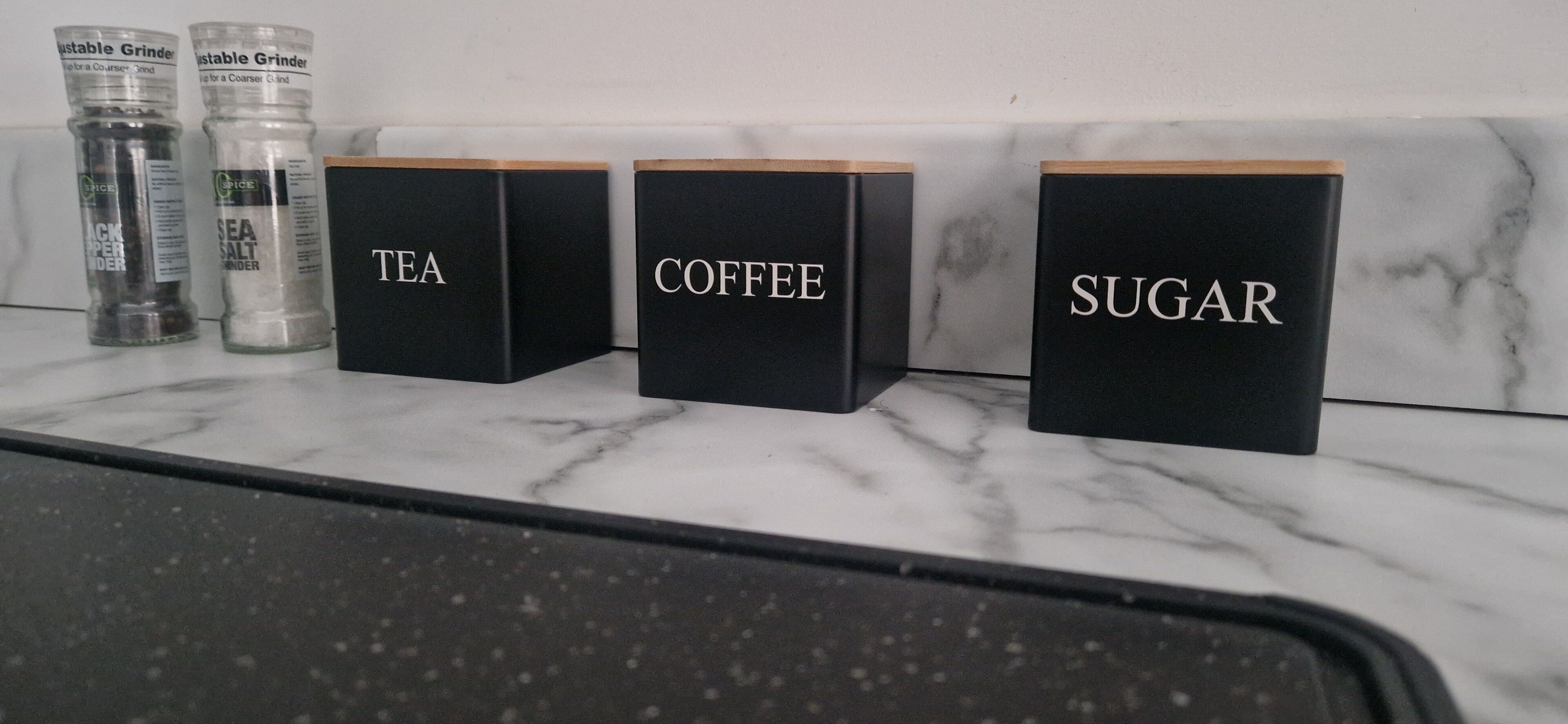 Three black containers labeled tea, coffee, and sugar on a marble countertop.
