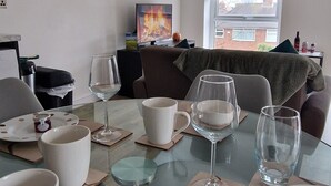 A modern living room with a glass dining table, a sofa, a TV, and a window with a view of a building. - Sarah's Woodland Golf Stays (Waterloo)