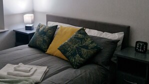 A neatly made bed with a gray mattress, a yellow and green patterned pillow, and a white pillow. There is a small bedside table with a lamp and a clock. The room has a plain white wall and a dark carpet. - Sarah's Woodland Golf Stays (Waterloo)
