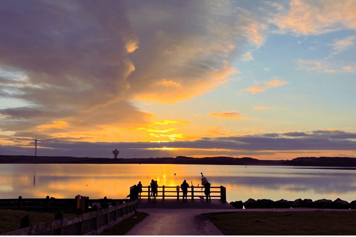 A sunset over a calm lake with a wooden pier and people standing at the end.