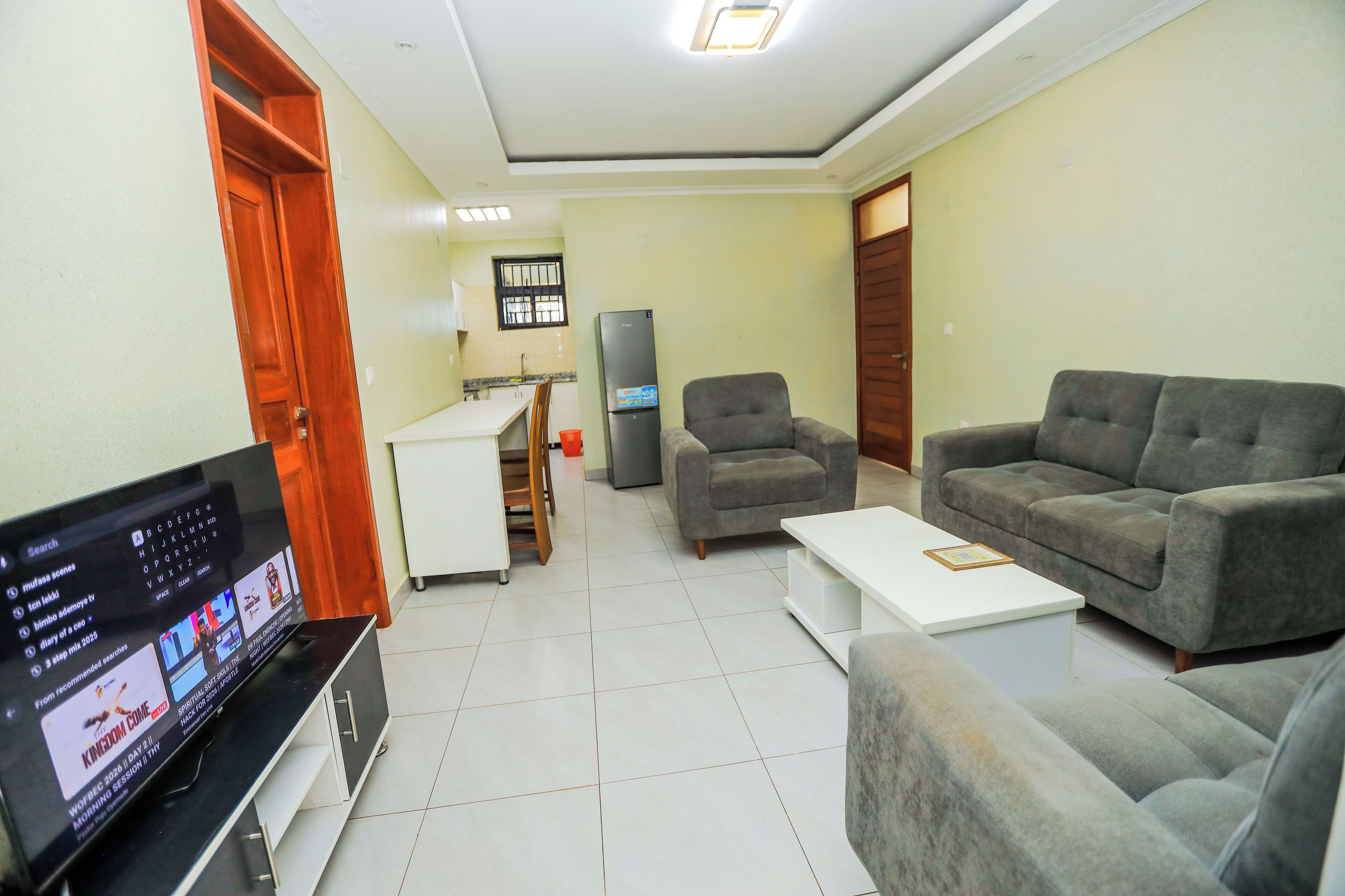 A living room with a flat-screen TV, a sofa, a coffee table, and a kitchen area in the background.