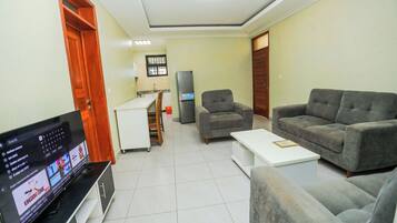 A living room with a flat-screen TV, a sofa, a coffee table, and a kitchen area in the background.
