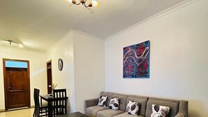 A living room with a sectional sofa, a coffee table, a dining table with chairs, a chandelier, and a painting on the wall. - ORVENA RESIDENCE (Kigali)