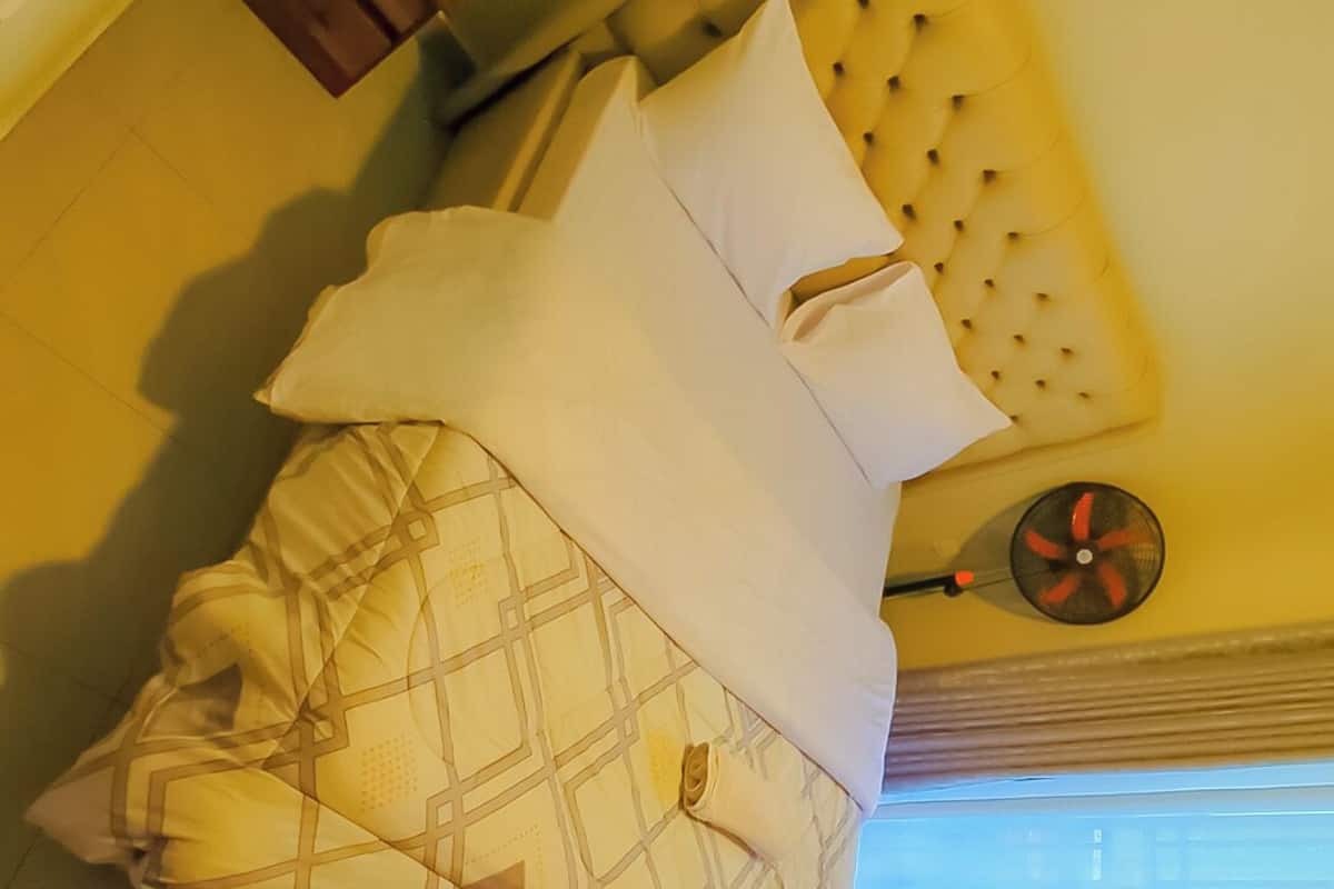 En seng med et mønstret tæppe og puder, et lille træskab og en vægventilator.