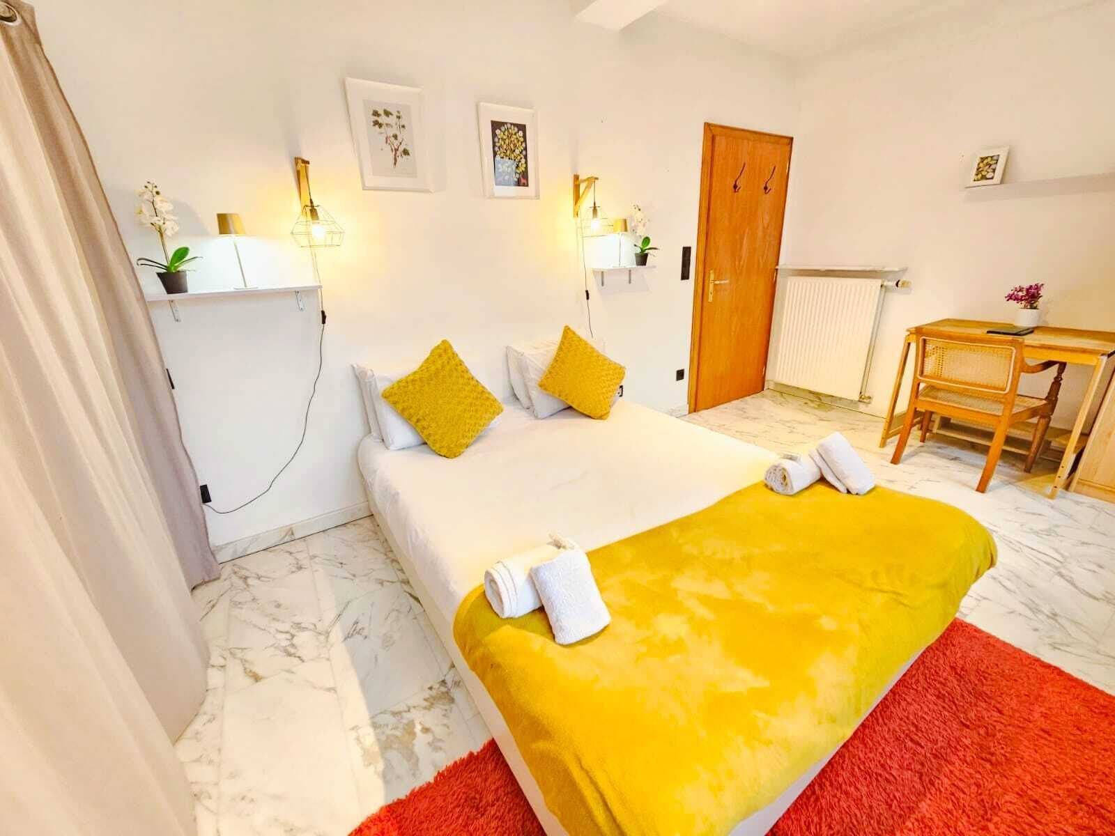 A hotel room with a bed, a desk, and a chair. There are two wall-mounted lamps, a wooden door, and a radiator.
