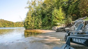 A lakeside setting with a sandy beach, a row of Adirondack chairs, and dense forest in the background. - Avec accès au Petit lac Long (Saint-Adolphe-d`Howard)