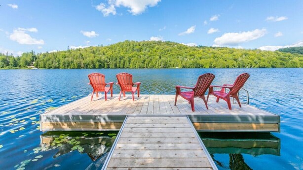 Een houten steiger met vier rode Adirondack-stoelen, gericht op een kalm meer, omringd door weelderige begroeiing en een helderblauwe lucht.