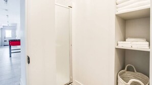 Een hotelkamer met een witte deur, een rode minibar en een plank met opgevouwen handdoeken en een gevlochten mand.