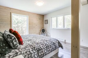 Un dormitorio con una cama, dos ventanas, una silla y suelo de madera.